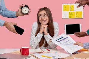 Pleased female entrepreneur keeps hands under chin, looks with gentle smile aside, poses ner unrecognizable people who hold alarm clock, touchpad, mobile phone, spiral notepad with stickers.