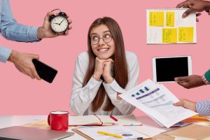Pleased female entrepreneur keeps hands under chin, looks with gentle smile aside, poses ner unrecognizable people who hold alarm clock, touchpad, mobile phone, spiral notepad with stickers.
