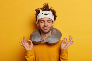 Relaxed passenger meditates indoor, tries to have rest during traveling in transport, wears neck pillow, has insomnia, stands in mudra gesture, poses against yellow wall. Airplane travel, comfort