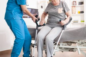 Nurse helping old woman to sit on bed in nursing home after walking with crutches.