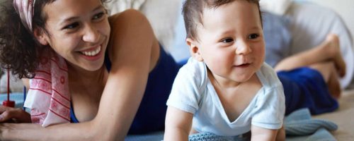 Childhood, physical development and childcare concept. Picture of attractive cheerful young mixed race female laughing, watching her adorable toddler child making first crawling movements on floor