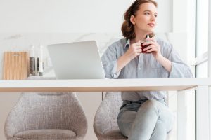 Portrait of cheerful woman in striped shirt and jeans holding cup of tea while sitting at soft chair in the kitchen