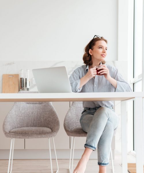 Portrait of cheerful woman in striped shirt and jeans holding cup of tea while sitting at soft chair in the kitchen