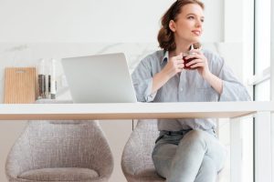 Portrait of cheerful woman in striped shirt and jeans holding cup of tea while sitting at soft chair in the kitchen
