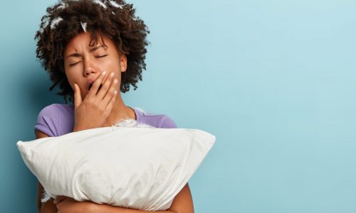 Sleepy tired young Afro American woman covers mouth with hand, yawns after sleep, embraces white pillow, has feathers in head, poses over blue background with blank space for your promotion.